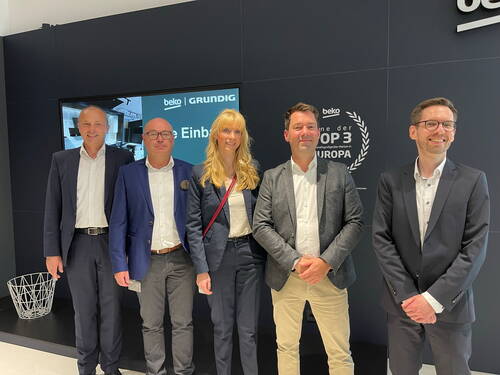 Fünf Geschäftsleute in Anzügen stehen lächelnd nebeneinander vor einer schwarzen Messewand mit den Markenlogos von Beko und Grundig.