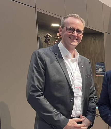 Lächelnder Mann im grauen Anzug mit Namensschild steht in modernem Messestand mit dunklen Schränken im Hintergrund