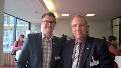 Zwei Geschäftsleute im Anzug lächeln in die Kamera, während sie bei einer Konferenz im Foyer mit Stehtischen und weiteren Teilnehmenden im Hintergrund stehen.