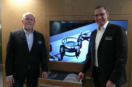Zwei Männer in Anzügen präsentieren in einer Ausstellungsküche einen geöffneten Geschirrspülauszug vor einem Monitor mit Großaufnahme eines modernen Gasherds.