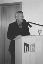 Älterer Mann im Anzug hält eine Rede an einem Rednerpult mit Mikrofon und hbc-Logo in einem Innenraum.