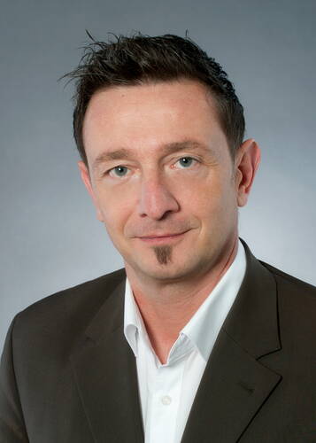 Mittelalterlicher Mann mit kurzem, dunkelbraunem Haar und kleinem Kinnbärtchen trägt weißen Hemdkragen und dunklen Anzug und blickt freundlich vor neutral grauem Hintergrund in die Kamera