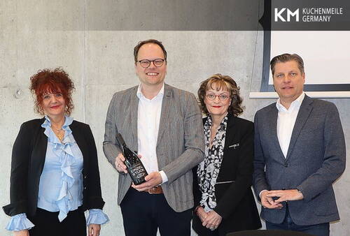 Vier Geschäftsleute stehen vor einer Betonwand, während der zweite von links lächelnd eine schwarze Auszeichnungstrophäe präsentiert, daneben das eingeblendete Logo „KM Küchenmeile Germany“.