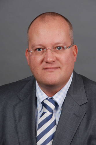 Glatzköpfiger Mann mit Brille im grauen Anzug und gestreifter Krawatte vor neutral grauem Hintergrund, der direkt in die Kamera blickt