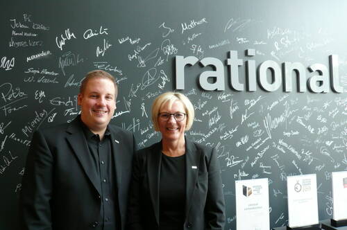 Zwei Geschäftsleute in schwarzen Anzügen lächeln vor einer signierten Wand mit dem Schriftzug „rational“ und mehreren Design-Auszeichnungen im Vordergrund