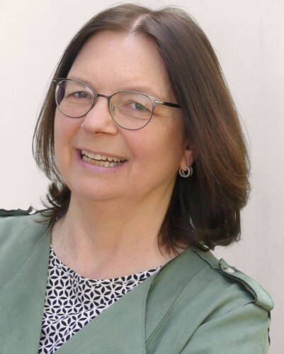 Ältere Frau mit schulterlangem braunem Haar und Brille lächelt freundlich in die Kamera, trägt eine grüne Jacke über einem gemusterten Oberteil vor hellem Hintergrund