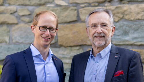 Zwei lächelnde Männer im Anzug stehen nebeneinander vor einer Natursteinwand und blicken direkt in die Kamera.