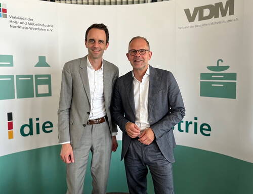 Zwei lächelnde Männer im Anzug posieren vor einem Messestand der Holz- und Möbelindustrieverbände Nordrhein-Westfalen und des Verbands der Deutschen Möbelindustrie.