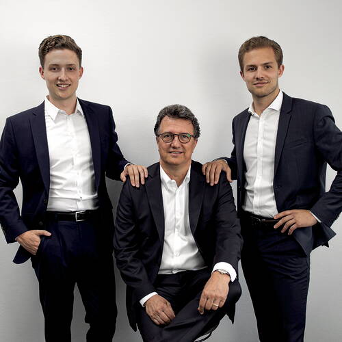 Drei Männer in dunklen Anzügen posieren vor einer weißen Wand, der ältere Mann sitzt in der Mitte auf einem Stuhl, während die beiden jüngeren links und rechts neben ihm stehen und ihm jeweils eine Hand unterstützend auf die Schulter legen.