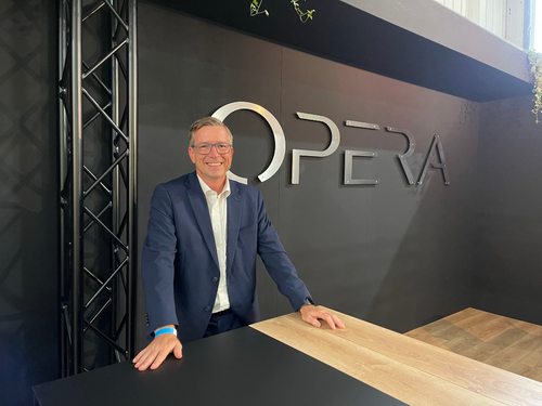 Lächelnder Mann im blauen Anzug steht an einem Holztresen vor einer dunklen Wand mit dem Schriftzug „OPERA“ in einem modernen Messestand.