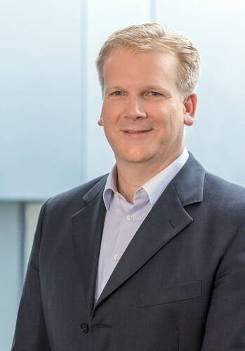 Mittelalterlicher Mann mit blondem, kurz geschnittenem Haar im dunklen Anzug und hellblauem Hemd lächelt in die Kamera vor einem hellen, unscharfen Hintergrund.