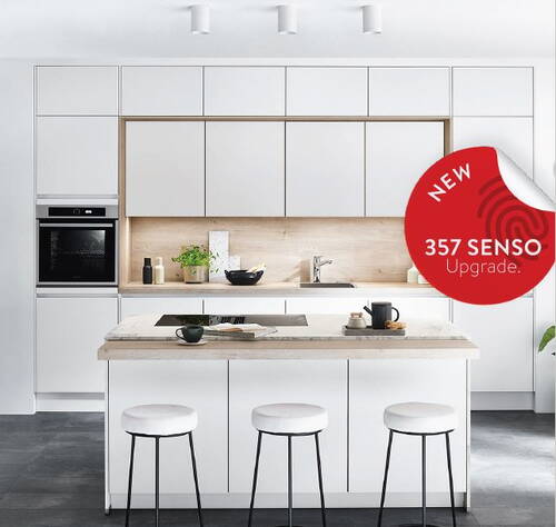 Helle, moderne Einbauküche in Weiß mit Kochinsel, drei Barhockern, Holzrückwand und rotem Hinweis auf das neue 357 SENSO Upgrade