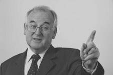Älterer Mann im Anzug spricht gestikulierend und hebt mahnend den Zeigefinger.
