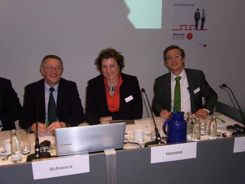 Drei lächelnde Konferenzteilnehmende sitzen hinter einem Tisch mit Laptop, Mikrofonen und Namensschildern in einem Tagungsraum.
