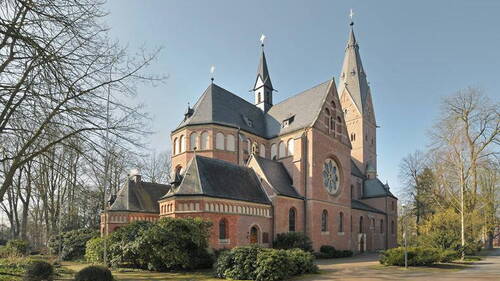 Backsteinkirche im neoromanischen Stil mit hohem Turm und Rosettenfenster, umgeben von Bäumen und gepflegten Grünanlagen an einem klaren, sonnigen Tag.