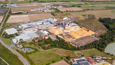 Großes Holzverarbeitungswerk mit weitläufigen Gebäuden, Sägespäne- und Holzlagerplätzen inmitten einer ländlichen Agrarlandschaft aus Feldern und Wiesen.