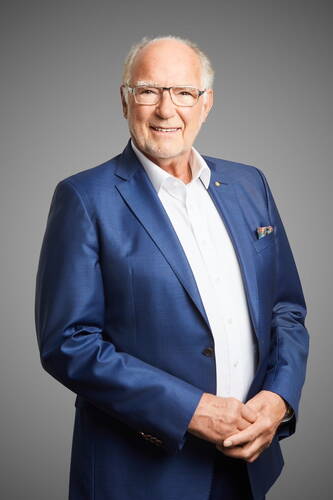 Älterer Mann mit Brille in elegantem blauen Anzug und weißem Hemd lächelt freundlich vor neutral grauem Hintergrund