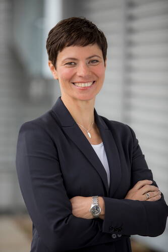 Selbstbewusste Geschäftsfrau mit Kurzhaarschnitt und dunklem Blazer steht mit verschränkten Armen lächelnd vor einem modernen, grauen Bürohintergrund