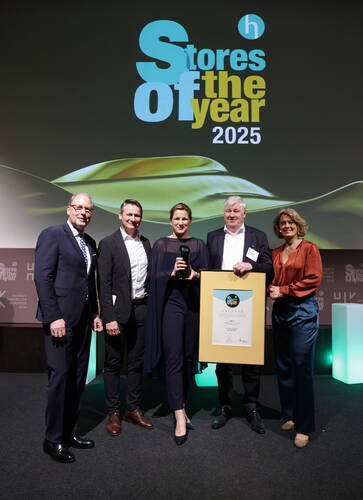 Fünf elegant gekleidete Personen posieren auf einer Bühne der „Stores of the Year 2025“-Preisverleihung, wobei eine Frau einen Glaspokal und ein Mann eine große gerahmte Urkunde in den Händen hält.