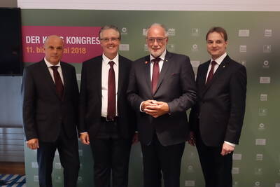 Vier Geschäftsleute in dunklen Anzügen posieren lächelnd vor einer Kongresswand mit der Aufschrift „Der Kog Kongress 11. bis 13. Mai 2018“.