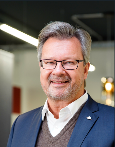 Älterer Mann mit Brille und grauem Haar lächelt im Business-Anzug in einem modern beleuchteten Büro.