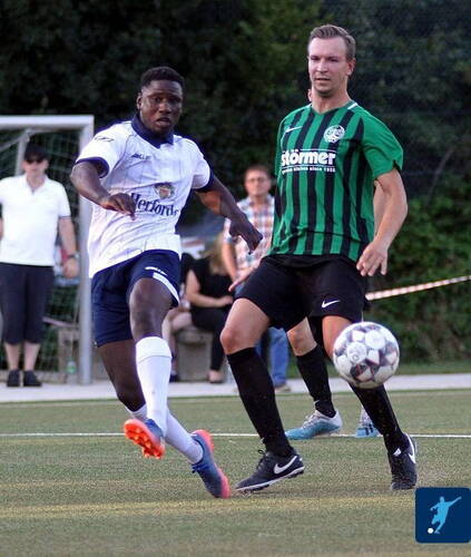 Zweikampf zweier Fußballer, einer im weißen und einer im grün-schwarz gestreiften Trikot, während der Spieler in Weiß den Ball im Lauf auf dem Kunstrasenplatz schießt und Zuschauer im Hintergrund zusehen.