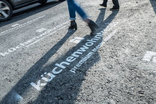 Personen mit Schatten laufen über eine grobe Asphaltfläche, auf der weiß der Schriftzug „küchenwohntrends premium messe“ als Wegmarkierung aufgesprüht ist.
