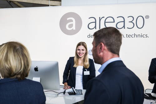 Freundliche Messemitarbeiterin begrüßt Besucher am Empfangsstand von area30 vor einem großen Markenlogo.