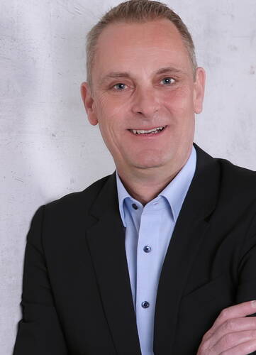 Lächelnder Mann im dunklen Anzug und hellblauem Hemd vor heller Betonwand, mit verschränkten Armen in einer professionellen Porträtaufnahme