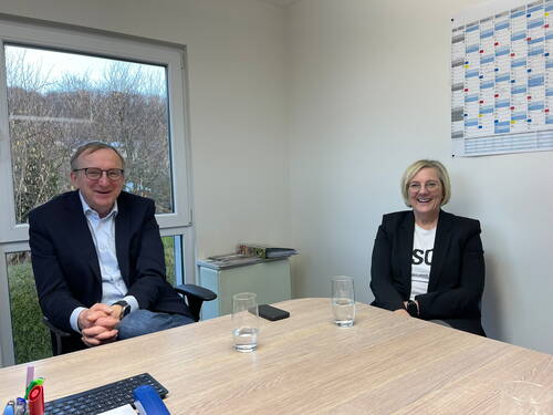 Zwei lächelnde Personen im Business-Outfit sitzen sich in einem hellen Büro gegenüber, vor einem Fenster mit winterlicher Baumlandschaft und einem großen Wandkalender im Hintergrund.