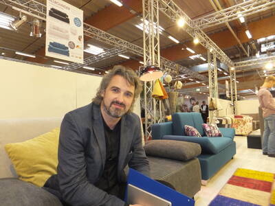 Mittelalterlicher Mann mit Bart sitzt lächelnd auf einem Sofa in einer modern beleuchteten Möbelmesse und hält eine blaue Mappe in den Händen.