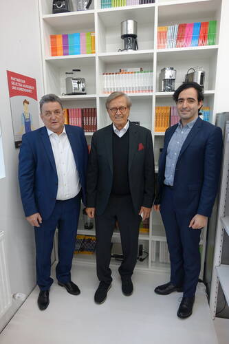 Drei Männer in Anzügen stehen lächelnd in einem modernen Showroom vor einem weißen Regal mit bunten Büchern und Küchenmaschinen
