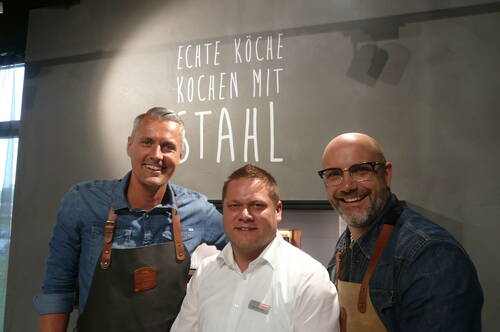 Drei lächelnde Männer, zwei mit Lederkochschürzen und einer in weißem Hemd, posieren in einer modernen Küche vor der Wandaufschrift „Echte Köche kochen mit Stahl“.