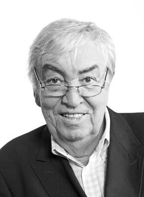 Älterer Mann mit grauen Haaren und Brille lächelt freundlich in die Kamera, elegant gekleidet im Sakko und Hemd vor hellem Hintergrund.