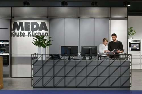 Zwei Mitarbeitende beraten sich hinter einem modernen Empfangstresen im Küchenstudio „MEDA Gute Küchen“ und blättern gemeinsam in einem Katalog.