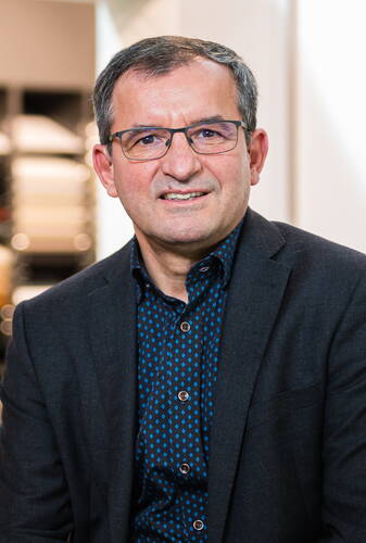Mittelalterlicher Mann mit Brille in dunklem Sakko und gemusteretem Hemd lächelt leicht in einer modernen Innenraumszene