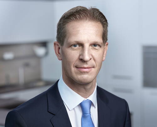 Geschäftsmann im dunklen Anzug mit blauer Krawatte blickt direkt in die Kamera in einem modernen, hellen Büro.