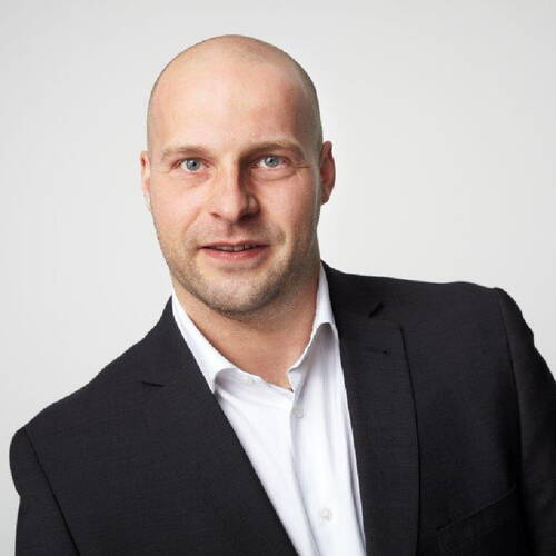 Glatzköpfiger Mann im dunklen Anzug und weißem Hemd lächelt leicht in einem professionellen Porträt vor hellem Hintergrund
