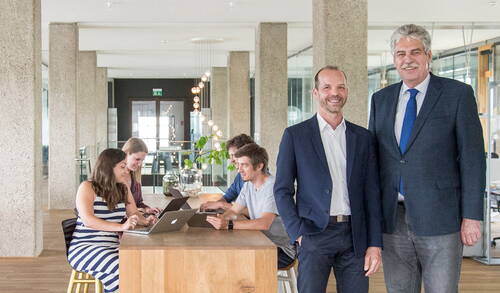 Zwei Männer im Anzug stehen lächelnd in einem modernen Großraumbüro, während ein gemischtes Team hinter ihnen gemeinsam an Laptops an einem hohen Holztisch arbeitet.