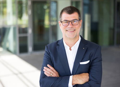Mittelalterlicher Mann im Anzug mit verschränkten Armen lächelt selbstbewusst vor einem modernen Bürogebäude