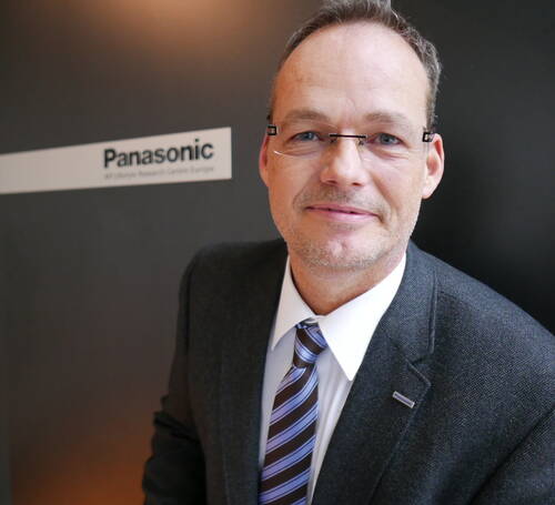 Geschäftsmann im Anzug mit gestreifter Krawatte lächelt in die Kamera, im Hintergrund ein Schild mit dem Panasonic‑Logo im Büro.