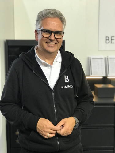 Älterer Mann mit Brille und grauen Haaren lächelt in schwarzer BELMENTO-Kapuzenjacke vor einem hellen Messestand.