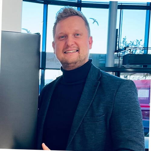 Lächelnder Mann im dunklen Rollkragenpullover und Sakko steht in einem modernen, lichtdurchfluteten Bürogebäude vor großen Glasfenstern.