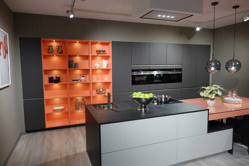 Modern eingerichtete Designküche mit grauen Schränken, offenem orangefarbenem Regal, zentraler Kochinsel mit Obstschale und Kochfeld sowie angrenzendem rosa Esstisch mit Blumenarrangement.