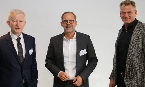Drei lächelnde Geschäftsleute in Anzug und Sakko posieren nebeneinander vor einer hellen, neutralen Wand bei einer Konferenz.