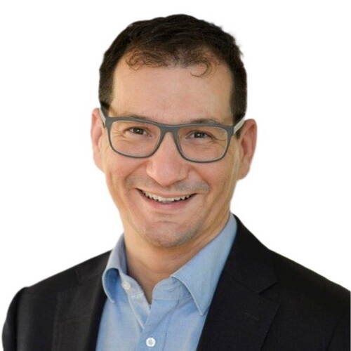 Lächelnder Mann mit Kurzhaarfrisur und Brille im dunklen Sakko und hellblauen Hemd vor weißem Hintergrund