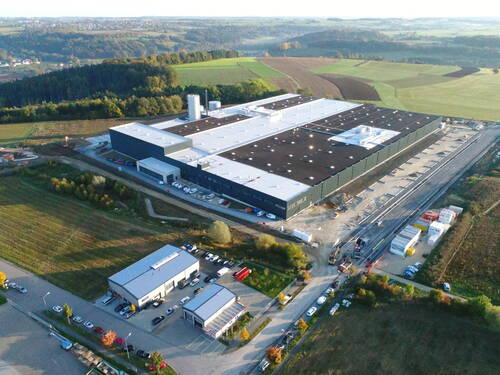 Großes, neu errichtetes Industrie- und Logistikgebäude am Rand einer ländlichen Ortschaft, umgeben von Feldern und Hügeln, mit Parkplätzen, Zufahrtsstraßen und noch laufenden Bauarbeiten auf dem Gelände