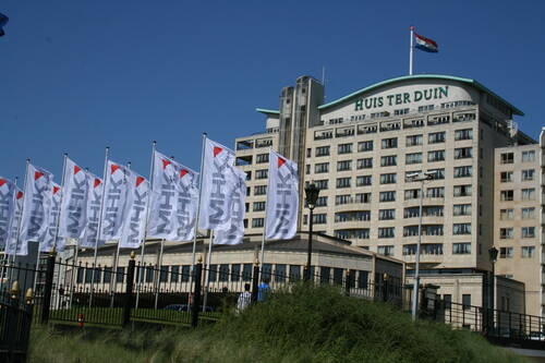 Mehrstöckiges Küstenhotel „Huis ter Duin“ mit niederländischer Flagge auf dem Dach und einer Reihe weißer MHK-Fahnen im Wind vor blauem Himmel.