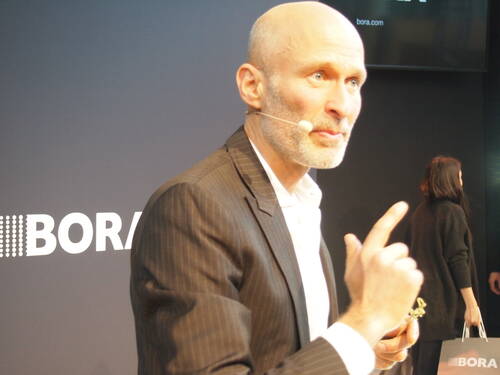 Kahlköpfiger Redner im Anzug mit Headset-Mikrofon gestikuliert während einer Präsentation auf einer Bühne mit BORA-Logo im Hintergrund.