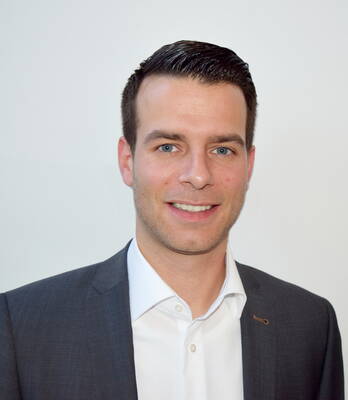 Junger Mann im dunklen Anzug und weißem Hemd lächelt freundlich vor einem hellen, neutralen Hintergrund